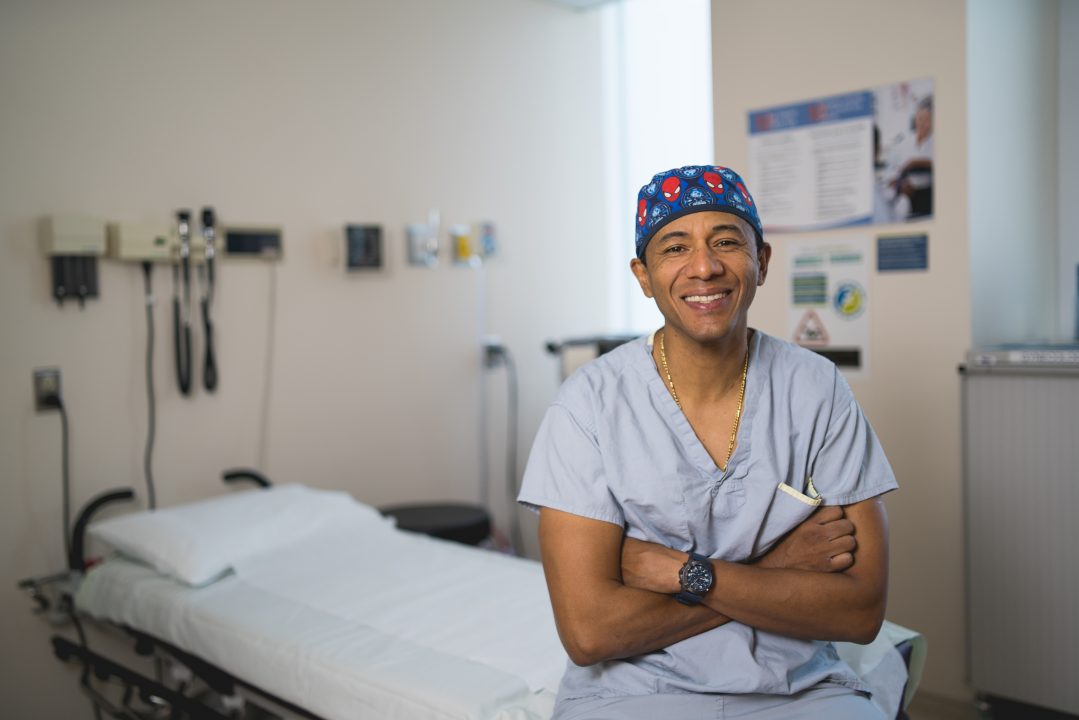 Dr. Antonio Caycedo, colorectal surgeon at Health Sciences North in Sudbury, Ontario, sits in a patient room, wearing surgical scrubs and a surgical scrub cap.